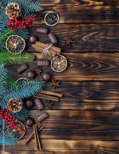 Chocolates, cinnamon tubes, star anise and dried lemons on a dark brown wooden background. Blue and green fir branches with cones and red berries in the background. Top view