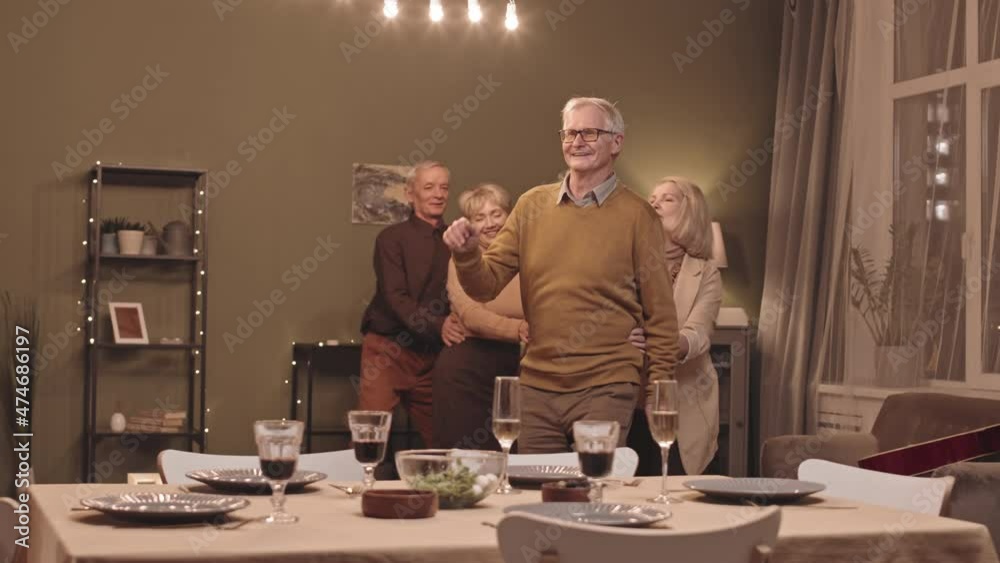Slowmo shot of five happy active seniors making a train and dancing together at home party in big cozy living room decorated with yellow lights