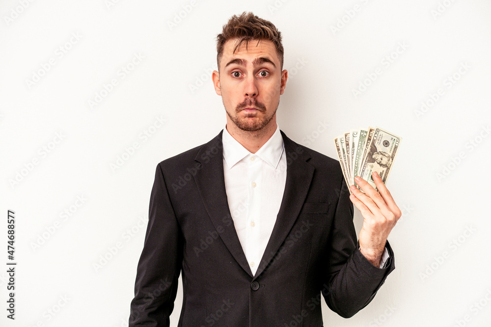 Young caucasian business man holding bank notes isolated on white background shrugs shoulders and open eyes confused. Young caucasian business man holding bank notes isolated on white background shrugs shoulders and open eyes confused.