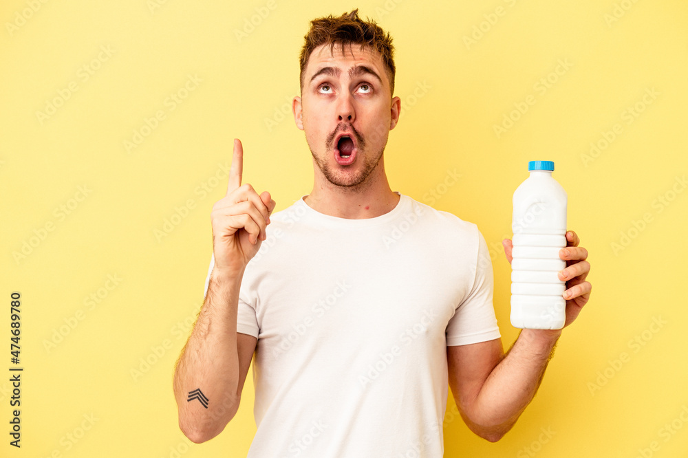 Young caucasian man holding a bottle of mil isolated on yellow background pointing upside with opened mouth.