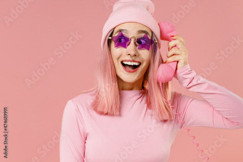 Young fun woman with bright dyed rose hair in rosy top shirt hat glasses hold handset vintage phone look aside isolated on plain light pastel pink background studio. People lifestyle fashion concept. © ViDi Studio