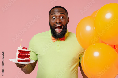 Young excited fun happy black gay man in green t-shirt bow tie hold bunch of air inflated helium balloons celebrating birthday party sweet cake with candle isolated on plain pastel pink background © ViDi Studio