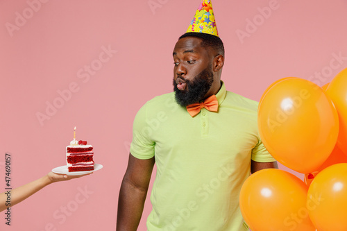 Young black gay man in green t-shirt hat cone hold bunch of air inflated helium balloons celebrate birthday party hand give cake with blow out candle isolated on plain pastel pink background studio. © ViDi Studio