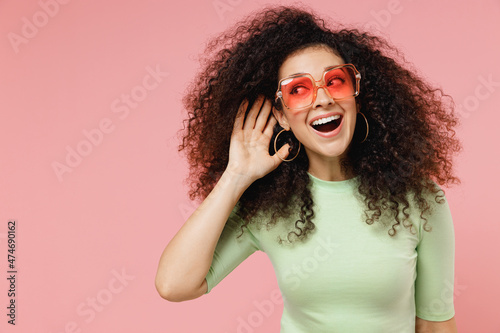 Curious nosy jubilant fun young curly latin woman 20s years old wear mint t-shirt sunglasses try to hear you overhear listening intently isolated on plain pastel light pink background studio portrait
