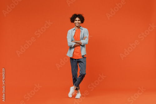 Full body young smiling black student man 50s wearing blue shirt t-shirt looking camera hold hands crossed folded isolated on plain orange color background studio portrait. People lifestyle concept © ViDi Studio