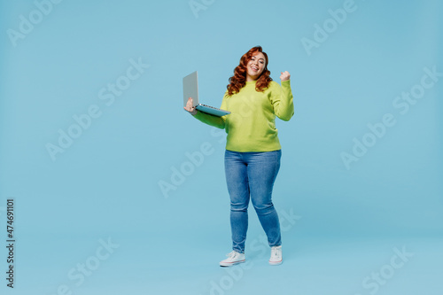 Full body smiling young chubby overweight plus size big fat fit woman in green sweater hold use work laptop pc computer do winner gesture isolated on plain blue background. People lifestyle concept