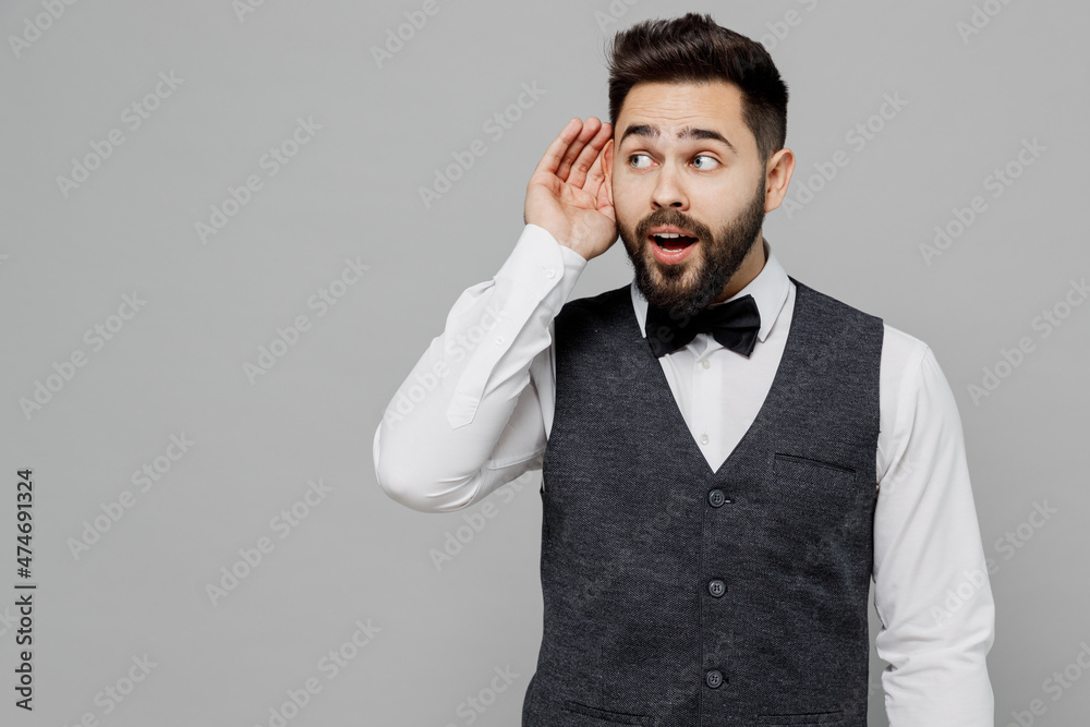 Young barista male waiter butler man 20s wear white shirt vest elegant uniform work at cafe try to hear you overhear listening intently isolated on plain grey background. Restaurant employee concept. Young barista male waiter butler man 20s wear white shirt vest elegant uniform work at cafe try to hear you overhear listening intently isolated on plain grey background. Restaurant employee concept.