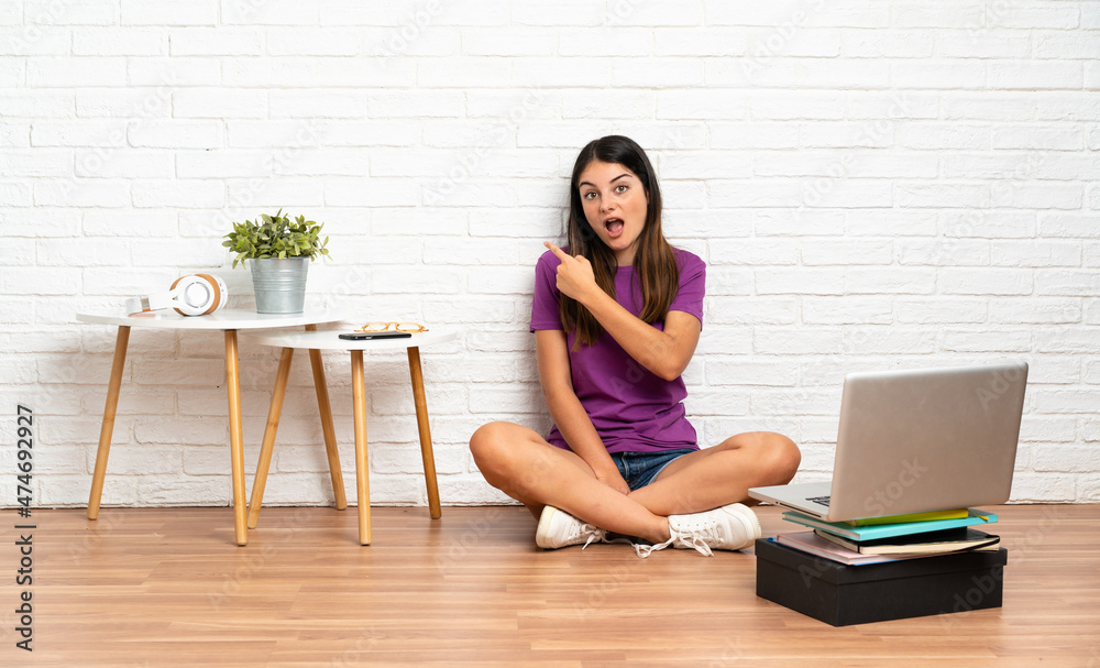 Young woman with a laptop sitting on the floor at indoors surprised and pointing side Young woman with a laptop sitting on the floor at indoors surprised and pointing side