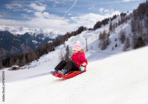 childhood, sledging and season concept - happy little girl sliding down on sled outdoors in winter over alps mountains on background