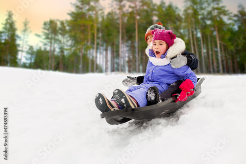 childhood, sledging and season concept - group of happy little kids sliding on sled down snow hill in winter over snowy forest or park background