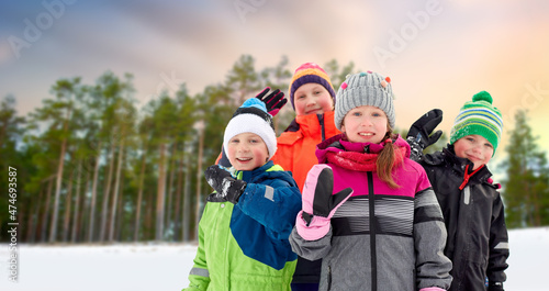 childhood, friendship and season concept - group of happy little kids in winter clothes outdoors over pine forest on background