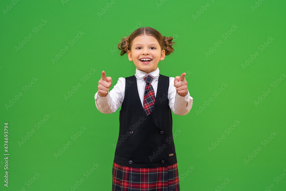 A smiling little schoolgirl points her index fingers forward. Isolated green background. A smiling little schoolgirl points her index fingers forward. Isolated green background.