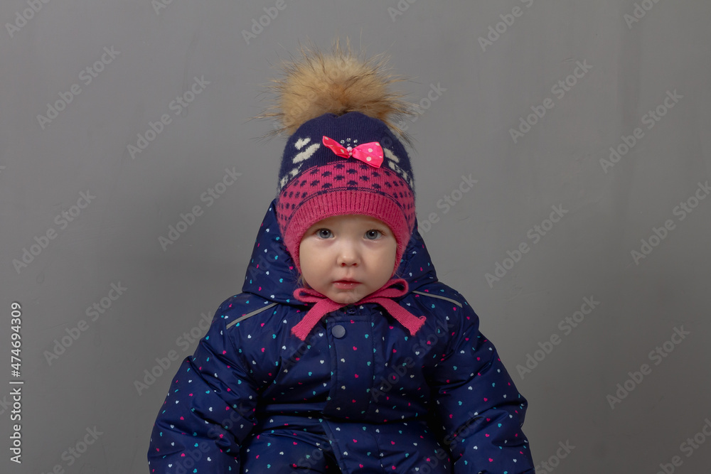 Cute baby girl 2 years old caucasian in warm winter clothes blue jumpsuit, hat, pink boots and mittens on a gray background
