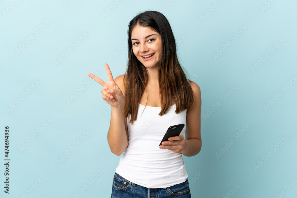 Young caucasian woman using mobile phone isolated on blue background smiling and showing victory sign Young caucasian woman using mobile phone isolated on blue background smiling and showing victory sign