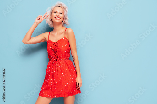 Portrait of young beautiful smiling female in trendy summer red dress. Sexy carefree woman posing near blue wall in studio. Positive blond model having fun and going crazy. Cheerful and happy