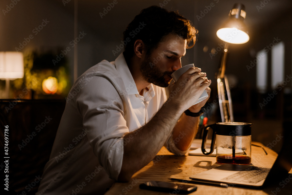 White bearded man drinking coffee while working with laptop in evening White bearded man drinking coffee while working with laptop in evening