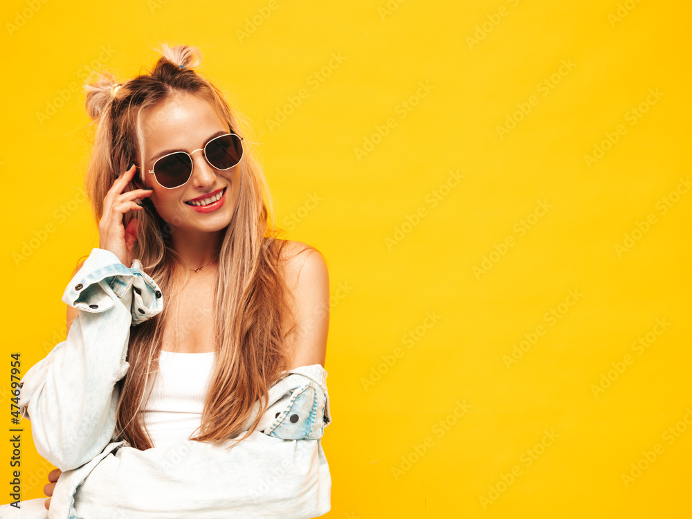 Fototapeta premium Portrait of young beautiful smiling blond female in trendy summer clothes. Sexy carefree woman posing near yellow wall in studio. Positive model having fun indoors. Cheerful and happy in sunglasses