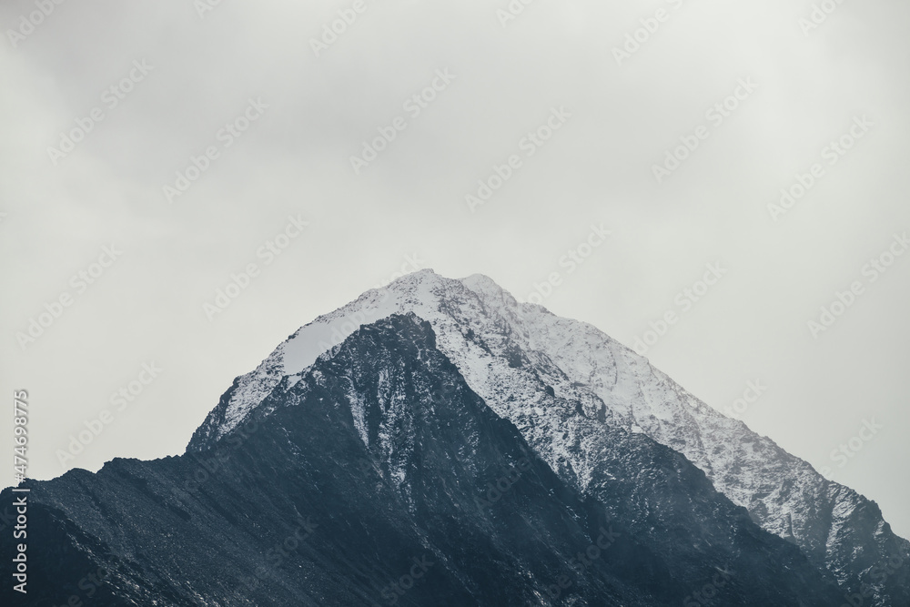 Dark atmospheric landscape with high black rocky mountain top with snow in cloudy sky. Dramatic mountain landscape with snow peaked top in haze in overcast weather. Gloomy view to awesome rocks in fog