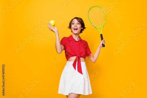 Portrait of excited pretty girl hold tennis ball racket look empty space open mouth smile isolated on yellow color background