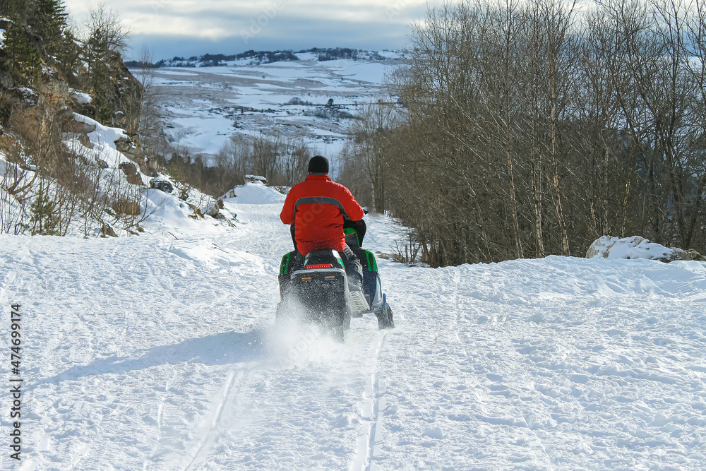A man in a red jacket is riding a snowmobile. In the background are snowy mountains and bare woods. It's a frosty, sunny day. Active recreation in winter.