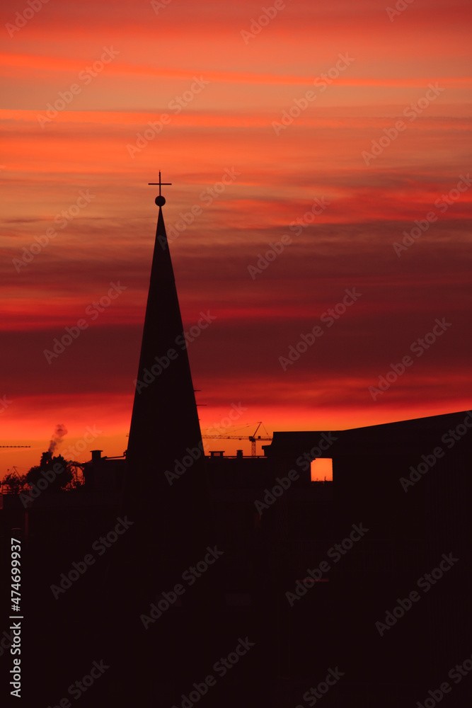 Urban winter sunset silhouette of the roofs of an italian city. Streaked burning red and orange sky, church steeple, see through lit windows. Cozy atmosphere, magic scenery. Golden hour.