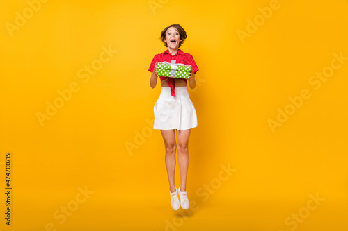 Full size photo of impressed nice brunette lady jump with present wear shirt skirt isolated on yellow background