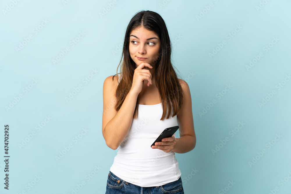 Young caucasian woman using mobile phone isolated on blue background having doubts and thinking Young caucasian woman using mobile phone isolated on blue background having doubts and thinking