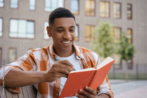 Smiling African American student studying, taking notes, exam preparation sitting in university campus. Education concept. Portrait young of confident writer planning project working outdoors 