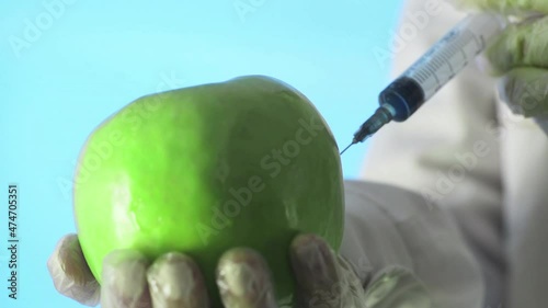 Close-up of a human in a medical coat and gloves injecting a syringe into an apple with some liquid on a blue background