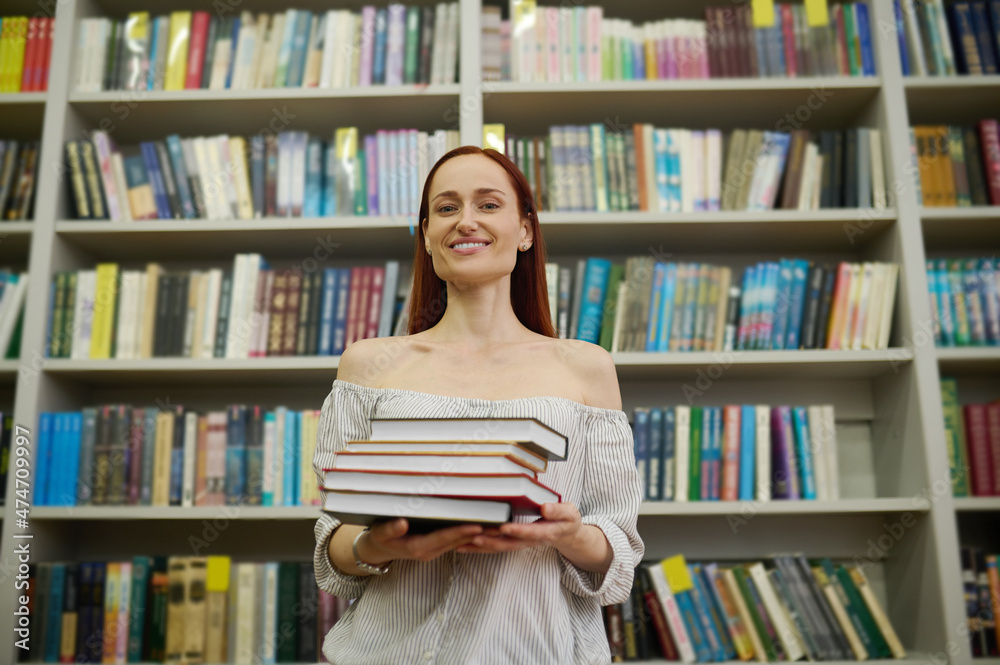Woman holding stack of books near bookshelves Woman holding stack of books near bookshelves