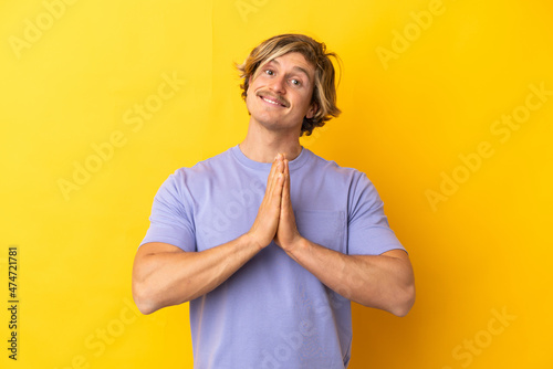 Handsome blonde man isolated on yellow background keeps palm together. Person asks for something