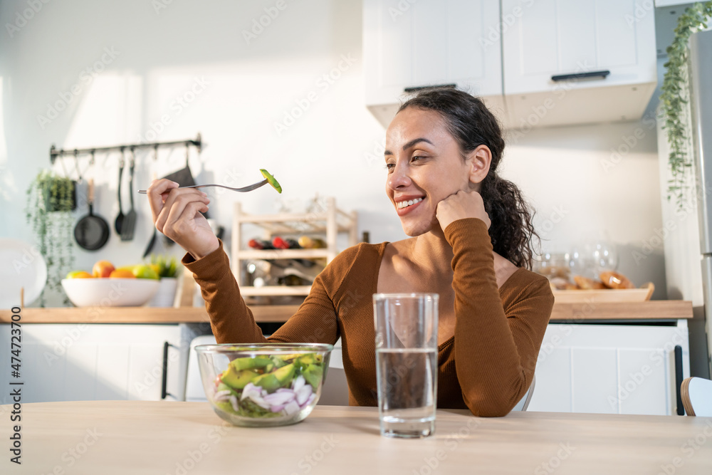 Young happy attractive Latino woman eat green salad in kitchen at home