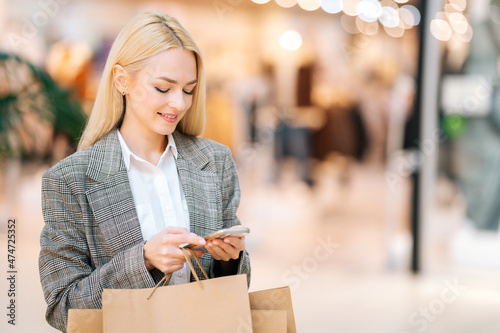 Portrait of smiling elegance blonde young woman in stylish jacket using mobile phone holding shopping paper bags with purchases in mall. Pretty shopaholic lady typing message on smartphone at store. © dikushin