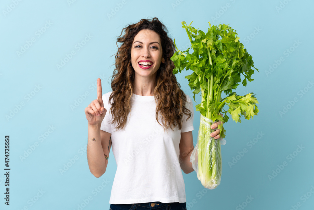Young woman holding a celery isolated on blue background pointing up a great idea Young woman holding a celery isolated on blue background pointing up a great idea