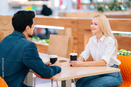 Happy handsome bearded man and attractive blonde woman having date sitting at table in mall, blurred background. Beautiful young couple drinking coffee and talking in cafe after shopping in store.