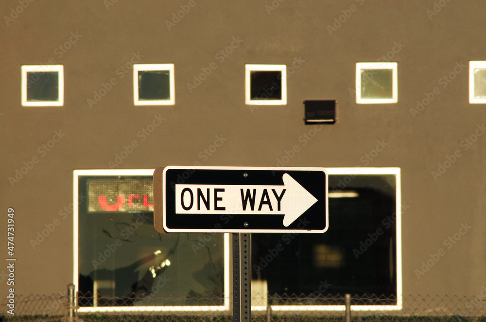 Obraz premium A one-way traffic sign is positioned in front of a commercial building facade with square windows and a lit 