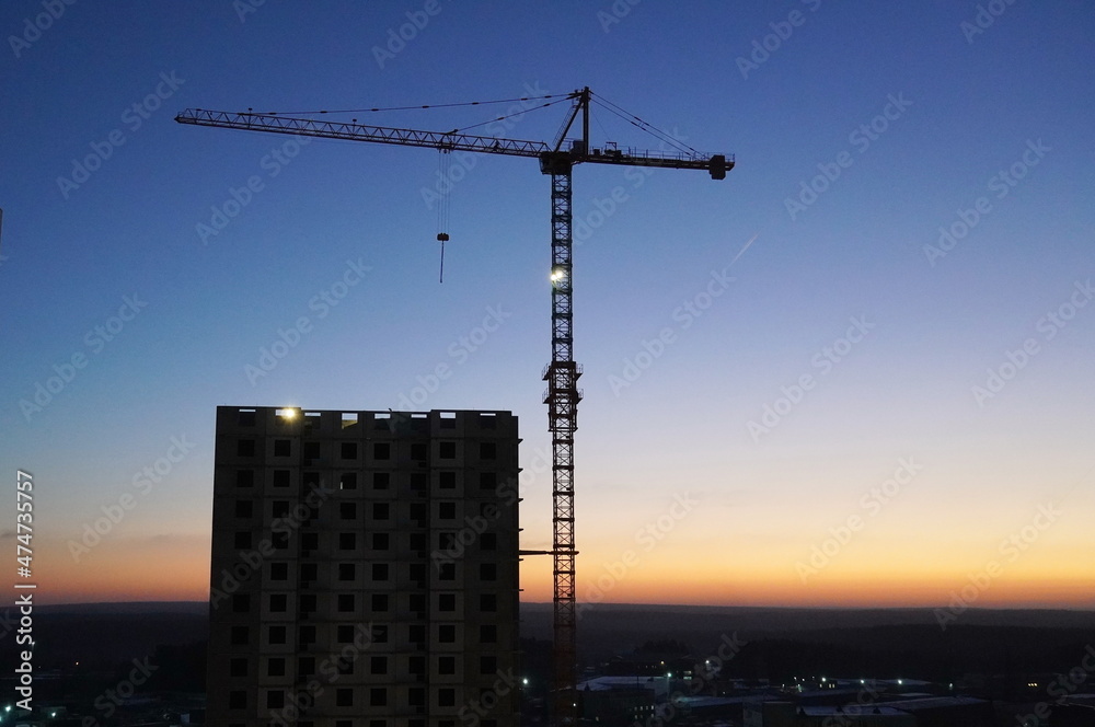 The dark silhouette of a construction crane and a multi-storey building in the early morning beautiful dawn. The dawn of a dark blue sky, a bright flaming sunny horizon and the silhouette of a crane.