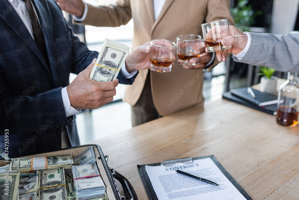 Fototapeta premium cropped view of businessman holding dollars while clinking glasses near contract and briefcase