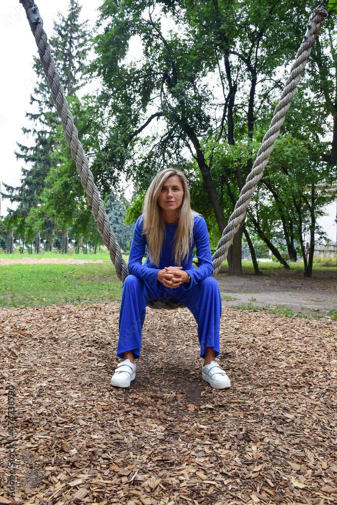 Fototapeta premium Attractive blonde woman sitting on a hanging rope swing in a blue suit in the park