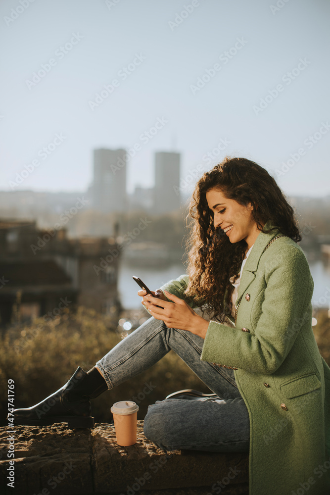 Pretty young woman using smartphone while sitting by the river and drinking takeaway coffee