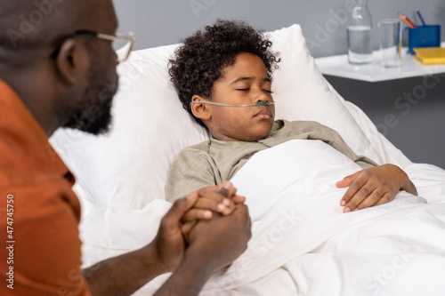 African little boy sleeping in bed at hospital ward with father holding his hand during the visit