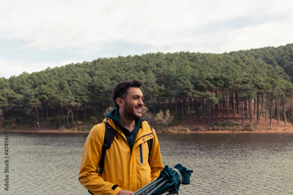 Fototapeta premium A boy with yellow raincoat standing in front of the lake. He has camp chair on his hand. He will have a rest by the lake. Nature park on a gold autumn day. He admires autumn nature landscape.