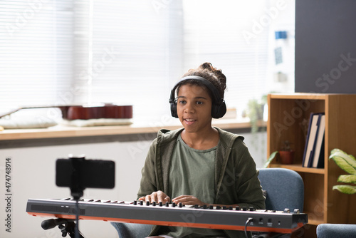 Happy teenage girl in headphones looking in smartphone camera while playing music keyboard in home environment