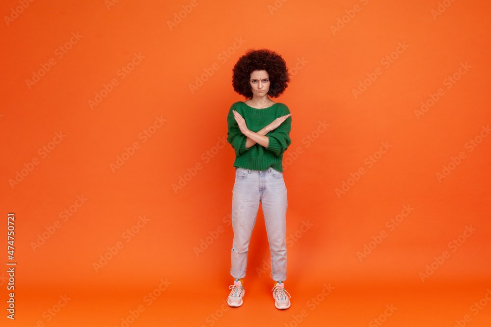 Finish, this is the end. Full length portrait of woman with Afro hairstyle wearing green sweater standing crossing hands, showing x sign meaning stop. Indoor studio shot isolated on orange background.