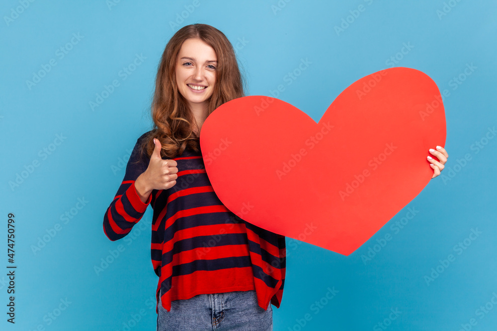 Positive woman wearing striped casual style sweater, holding big red heart, symbol of love and showing thumb up, expressing romantic feelings. Indoor studio shot isolated on blue background. Positive woman wearing striped casual style sweater, holding big red heart, symbol of love and showing thumb up, expressing romantic feelings. Indoor studio shot isolated on blue background.