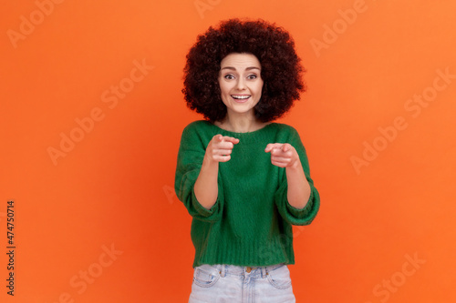 Happy positive woman with Afro hairstyle wearing green casual style sweater pointing finger to camera with happy expression, choosing you. Indoor studio shot isolated on orange background.