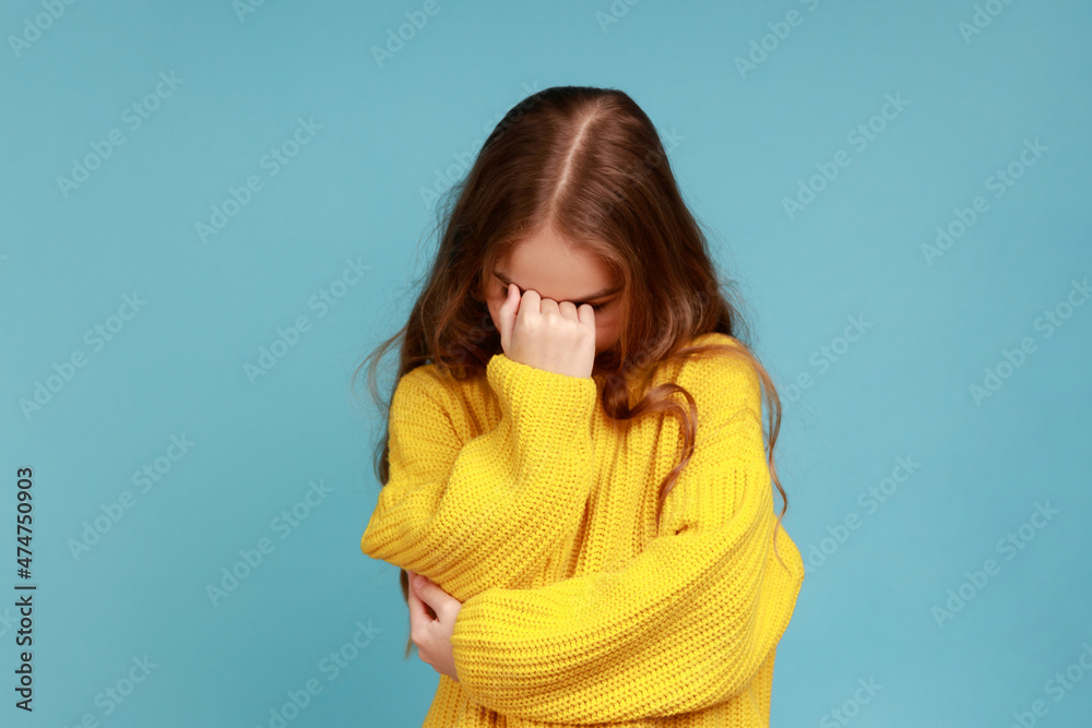 Portrait of little girl hiding face down and crying, upset about loss, feeling sorrow and regret, wearing yellow casual style sweater. Indoor studio shot isolated on blue background. Portrait of little girl hiding face down and crying, upset about loss, feeling sorrow and regret, wearing yellow casual style sweater. Indoor studio shot isolated on blue background.