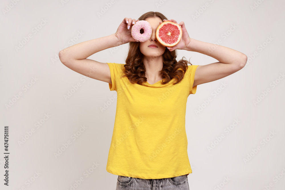 Sugary dessert vs fruits. Portrait of playful brown haired woman in yellow T-shirt covering eyes with fresh grapefruit and sweet doughnut, junk food. Indoor studio shot isolated on gray background. Sugary dessert vs fruits. Portrait of playful brown haired woman in yellow T-shirt covering eyes with fresh grapefruit and sweet doughnut, junk food. Indoor studio shot isolated on gray background.