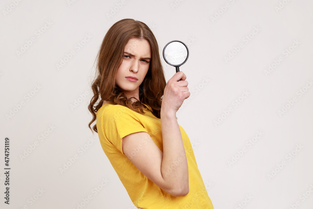 Serious confident teenager girl detective in casual style yellow t-shirt holding in hand magnifying glass and looking at camera, inspecting. Indoor studio shot isolated on gray background. Serious confident teenager girl detective in casual style yellow t-shirt holding in hand magnifying glass and looking at camera, inspecting. Indoor studio shot isolated on gray background.