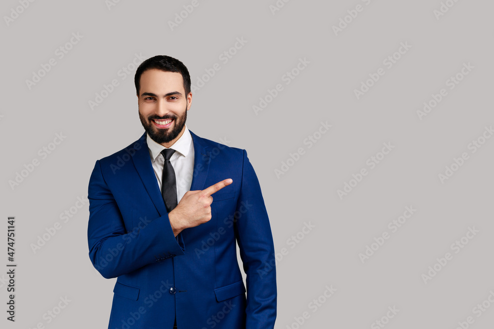 Positive bearded man pointing finger away with toothy smile on face, empty space for your advertisement, wearing official style suit. Indoor studio shot isolated on gray background. Positive bearded man pointing finger away with toothy smile on face, empty space for your advertisement, wearing official style suit. Indoor studio shot isolated on gray background.
